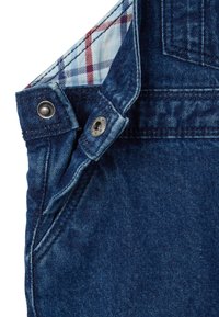 Los overoles de mezclilla presentan una tela azul oscuro con costuras visibles, detalles metálicos y un forro a cuadros en tonos de azul y rojo.