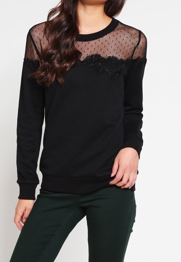 Femme portant un haut noir à manches longues avec un empiècement transparent à pois et un détail en dentelle florale, associé à un pantalon vert foncé.