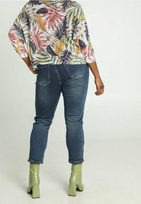 Blusa holgada con estampado floral y mangas tres cuartos, combinada con vaqueros ajustados azules y botines de tacón verdes. Detalles visibles de tela texturizada.