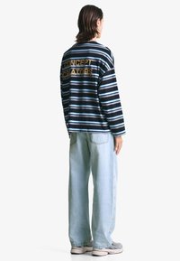 Randig långärmad skjorta i marinblått och ljusblått, med "CONCEPT CREATORS" i guld på baksidan; i kombination med ljusblå vida jeans.