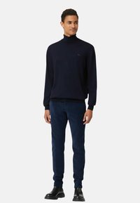 Maglione a dolcevita blu navy con maniche lunghe e polsini a costine, abbinato a pantaloni in velluto a coste scuro e stivaletti neri alla caviglia. Dettaglio logo semplice.