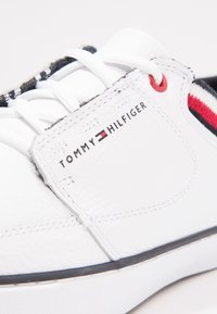 Baskets en cuir blanc à fini texturé, avec un détail à rayures noires et rouges, des lacets blancs et une écusson logo sur le côté.