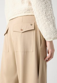 Beige tailored trousers with two rear pockets, button closures, and a smooth texture. The garment features a high waist and subtle pleats.