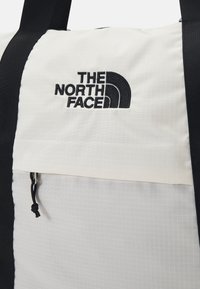 Fehér és fekete szövet sporttáska rácsos mintával, tetején cipzáras zseb, állítható pánttal és fekete hímezett The North Face logóval.