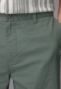 Pantaloni in tessuto verde con una texture liscia, dotati di tasche laterali, chiusura con bottone e dettagli di cucitura in vista.
