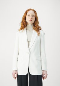 Blazer bianco con rever a punta, chiusura a un bottone e due tasche frontali, indossato sopra un dolcevita beige chiaro. Texture liscia.