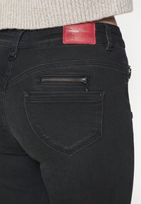 Jeans de mezclilla negra con una etiqueta de cuero roja en la cintura y un pequeño bolsillo con cremallera en la parte trasera, con una textura suave y un ajuste entallado.