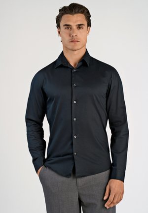 Uomo con indosso una camicia nera attillata a maniche lunghe abbottonata e pantaloni grigi, in piedi con una mano in tasca contro uno sfondo semplice.