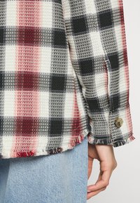 Gros plan d'une main à côté d'un ourlet effiloché d'une manche de chemise à carreaux rouge, noir et blanc, associée à un jean en denim bleu clair.