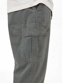 Vista lateral de pantalones cargo grises que muestran un gran bolsillo con solapa en el muslo, usados con una camisa de color claro.