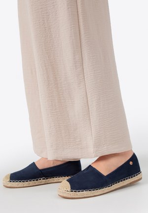 Pies calzados con alpargatas azul marino con punteras de tejido beige, combinadas con pantalones anchos de pierna texturizados en beige.