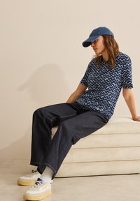Námořnická košile s krátkým rukávem a zvlněným vzorem, spárovaná s tmavými širokými kalhotami a béžovými teniskami. Modelka má na hlavě modrou kšiltovku.
