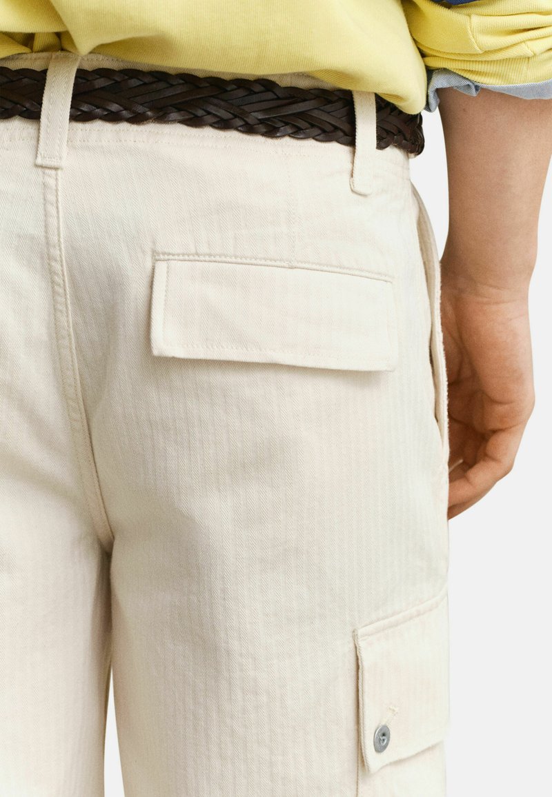 Gros plan sur une personne portant un pantalon cargo crème avec des poches à rabat, une ceinture tressée marron, et un haut jaune à manches longues retroussées.