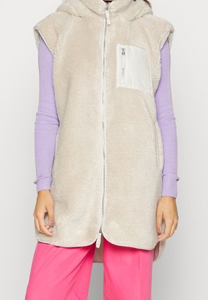 Beige faux fur vest med hætte, frontlynlås og en kontrastfarvet hvidlappetaske. Stylet over en lavendelribbet bluse med lange ærmer og pink bukser.