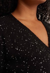 Črn pulover z v-izrezom, izdelan iz teksturirane tkanine, okrašen s srebrnimi bleščicami v cvetličnem vzorcu, ki poudarja sijoče detajle.