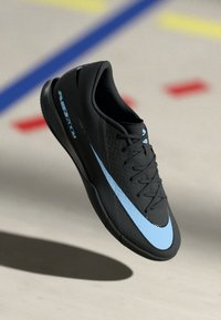 Zwarte sportieve schoen met een gestructureerde bovenkant, voorzien van een lichtblauwe swoosh-ontwerp en zwarte veters, weergegeven in een zwevende positie.