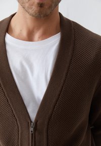 Braune Strickjacke mit strukturiertem Muster und silbernem Reißverschluss, über einem weißen T-Shirt mit Rundhalsausschnitt getragen. Strukturierte Stoffdetails sichtbar.