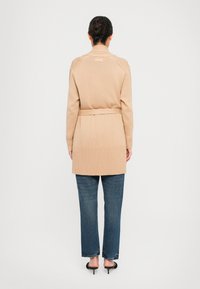 Nainen seisoo selkä kameraan, päällä beige vyöllinen neuletakki, siniset farkut ja mustat pikkukorkokengät tasaisella taustalla.