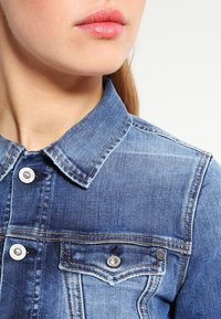Denim jacket featuring a light blue hue, traditional collar, two chest pockets with silver buttons, and contrast stitching. Textured fabric visible.