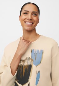 Beżowy sweatshirt z okrągłym dekoltem, zdobiony abstrakcyjnym nadrukiem w odcieniach niebieskiego, czarnego i złotego. Teksturowana tkanina, luźny krój i krótkie rękawy.
