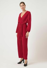 Rød plisseret jumpsuit med en dyb V-hals, brede ærmer og et matchende bælt. Lavet af glat stof, sat sammen med sorte højhælede sko.