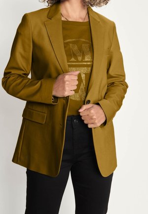 Kvinde iført olivengrøn blazer over matchende pyntet top og sorte bukser, justerer blazeren med begge hænder.