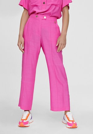 Kvinde iført højttaljede, lyserøde, brede bukser, stående med afslappede hænder ned langs siderne, kombineret med hvide, pink, orange og lilla sneakers.