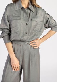 Šedá saténová košile s volným střihem, velkým límcem a dvěma předními kapsami. Disponuje zapínáním na knoflíky a ohrnutými rukávy, doplněná o odpovídající kalhoty.