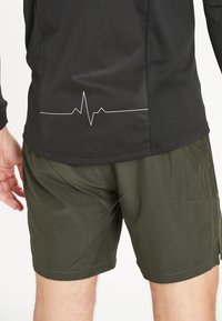 Zwart sportshirt met een hartslaglijn graphic op de achterkant, gecombineerd met olijfgroene shorts in een relaxte pasvorm.