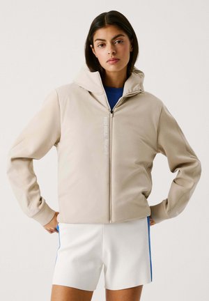 Beige zip-up hættetrøje med blød tekstur. Har "RED BULL RACING" branding. Bæres over en blå skjorte og hvide shorts med blå detaljer.