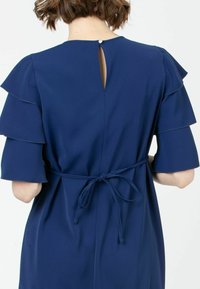 Laivansininen mekko, jossa on kerrokselliset röyhelöhihat, avotukka nappikiinnityksellä ja vyötärölle sidottu yksityiskohta, joka luo istuvan siluetin.