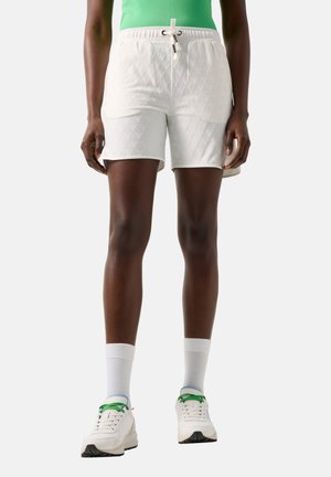 Person wearing white textured shorts with drawstring, white socks, white sneakers with green laces, and a green top, standing against a plain background.