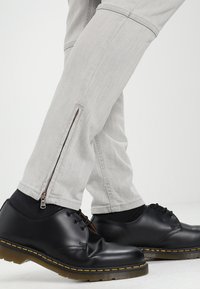 Musta nahkakengät, joissa on nauhat ja keltaiset saumat, yhdistetty vaaleanharmaisiin housuihin, joissa nilkoissa vetoketjut ja rento istuvuus.