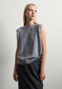 Femme aux longs cheveux blonds portant un pull en tricot gris sans manches et une jupe noire, se tenant contre un mur clair, regardant sur le côté.