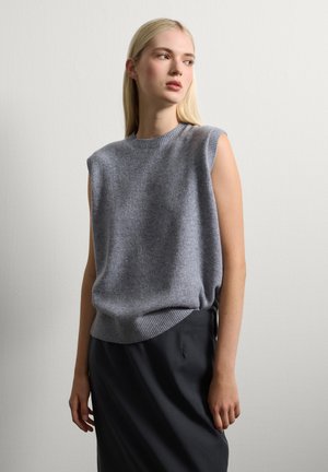 Femme aux longs cheveux blonds portant un pull en tricot gris sans manches et une jupe noire, se tenant contre un mur clair, regardant sur le côté.