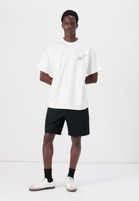 Hvit bomull T-skjorte med korte ermer og rund hals, med en liten grafisk detalj. Sammen med svarte shorts og hvite joggesko.