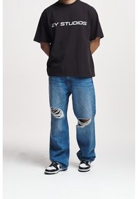 Person wearing a black "2Y STUDIOS" t-shirt, ripped blue jeans, and black-and-white sneakers, standing with hands behind back.