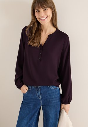 Blusa a maniche lunghe di un profondo viola con scollatura a V e pattino con bottoni; abbinata a jeans larghi blu. Tessuto morbido e vestibilità comoda.
