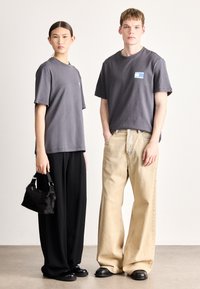 Dois modelos vestem t-shirts largas de algodão cinzentas com detalhes em remendo, combinadas com calças pretas de perna larga e jeans bege. Os acessórios incluem uma pequena bolsa preta.