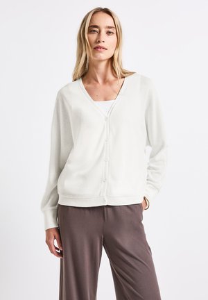 Weißer Cardigan mit V-Ausschnitt, vorderer Knopfleiste und langen Ärmeln, kombiniert mit braunen Hosen. Der Stoff scheint weich und strukturiert zu sein.