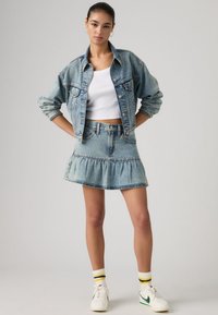 Συνδυασμός ανοιχτού μπλε τζιν μπουφάν και φούστας με βολάν στο τελείωμα. Λευκό φανελάκι, λευκά αθλητικά παπούτσια με πράσινες λεπτομέρειες και ριγέ κάλτσες.