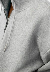 Sudadera con capucha gris con cremallera, hecha de tela suave; cuenta con una cremallera larga, cordones y una superficie texturizada, adecuada para ropa casual.