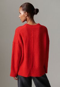 Röd oversized tröja i mjukt material, med ribbad halsringning och muddar, något längre bakre fåll och sänkt axel.