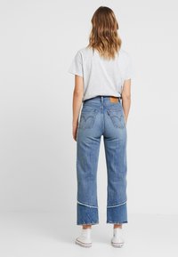 Lysegrå t-shirt og blå højtaljede jeans med frynset kant. Jeans har baglommer med karakteristisk syning og mærkelogo påsat.