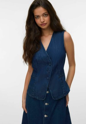 Femeie cu păr brunet lung și ondulat, purtând o vestă fără mâneci din denim albastru închis și o fustă asortată cu nasturi, pe un fundal simplu și deschis la culoare.