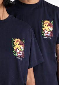 Marineblå bomulds T-shirts med et levende blomster- og botanisk trykdesign på venstre bryst, der viser teksten "L'Antidote."