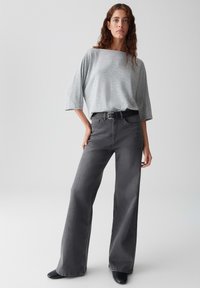 Femme aux cheveux bouclés portant un haut gris loose, un jean large gris foncé, une ceinture noire et des chaussures noires, se tenant devant un fond blanc uni.