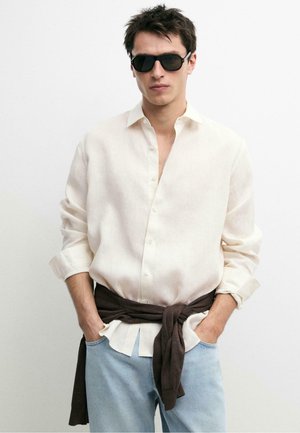 Mężczyzna w czarnych okularach przeciwsłonecznych, jasnobeżowej koszuli zapinanej na guziki, jasnoniebieskich dżinsach i ciemnym swetrze związanym w pasie, ręce w kieszeniach.