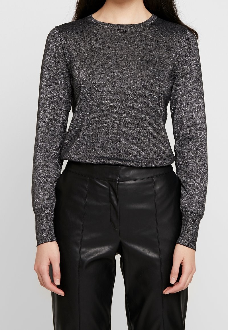 Kvinna som bär en metallisk långärmad topp instoppad i höga svarta läderbyxor, står med avslappnade armar längs sidorna.