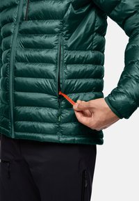 Giacca isolante verde con design trapuntato, dotata di una tasca con zip frontale e un brillante gancio arancione etichettato 'PACK AWAY'. Pantaloni neri in vista.